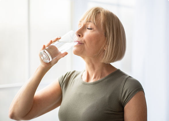 Mulher loira de perfil bebendo um copo de água com os olhos fechados.